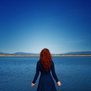 Fotografie Redhead in blue dress faces rippled lake, Anna Gorin, (40 x 40 cm)